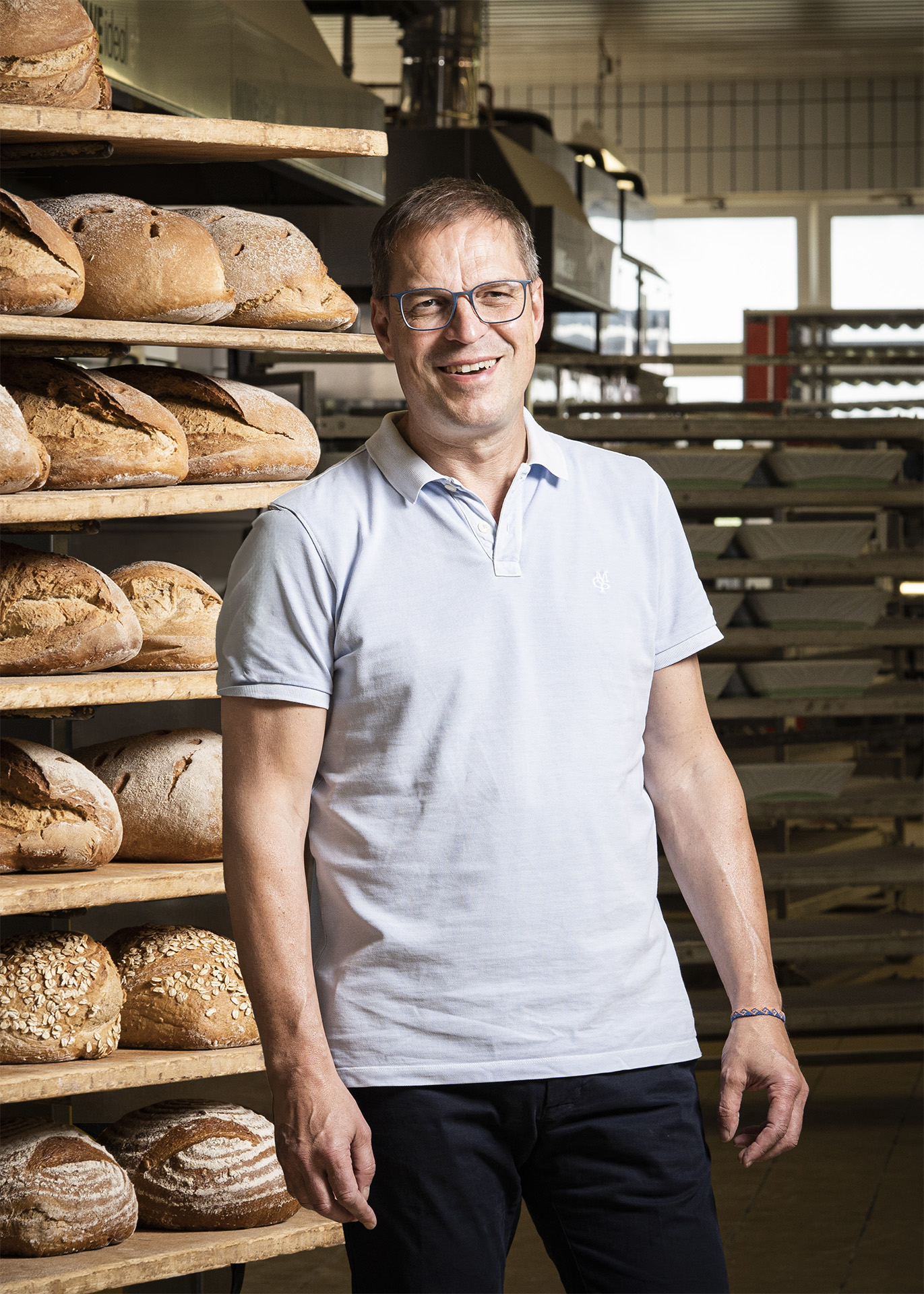 langs-backstube-baeckerei-ansprechpartner-joachim-lang_hoch Ein Mann mit Brille und Polohemd steht lächelnd in einer Backstube vor einem Regal voller Brot.