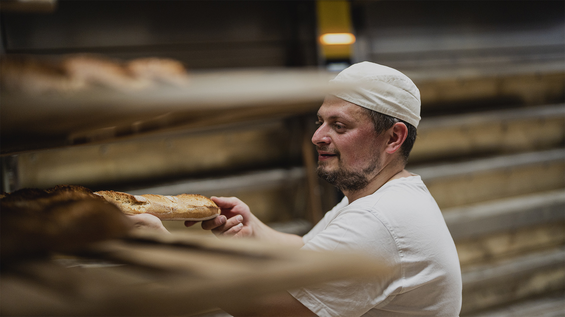 Bäcker mit weißer Mütze holt frisch gebackenes Baguette aus dem Ofenregal in der Bäckerei.